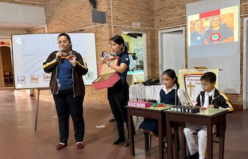 Laura Cubilla con gafas y micrófono, dirigiendo a alumnos en una actividad educativa. Niña y niño participan enfocados en materiales coloridos.