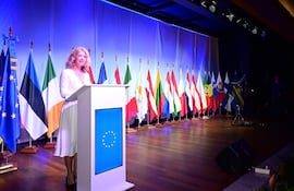 Mujer de cabello rizado y rubio en traje blanco habla en un atril frente a banderas de la UE y otros países.
