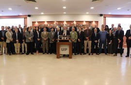 Hombre hablando frente a micrófono, rodeado de 40 personas en una sala formal, todos vestidos adecuadamente para la ocasión.