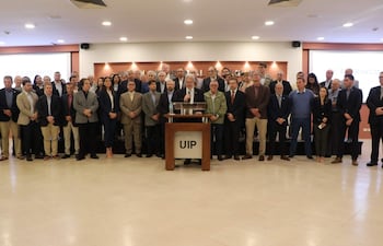 Hombre hablando frente a micrófono, rodeado de 40 personas en una sala formal, todos vestidos adecuadamente para la ocasión.