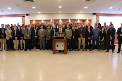 Hombre hablando frente a micrófono, rodeado de 40 personas en una sala formal, todos vestidos adecuadamente para la ocasión.