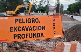 Cartel de advertencia naranja sobre excavación profunda y excavadora JCB en un sitio de construcción en zona urbana.