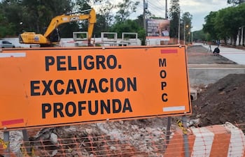 Cartel de advertencia naranja sobre excavación profunda y excavadora JCB en un sitio de construcción en zona urbana.