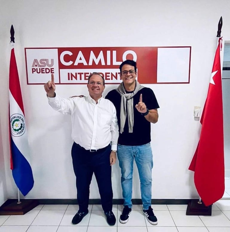 Camilo Pérez con camisa blanca y Nelson Mora en camiseta negra, ambos sonriendo y levantando el dedo índice en un ambiente de oficina.