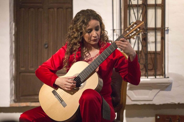 Mujer de cabello rizado, vestida de rojo, tocando guitarra acústica en un ambiente acogedor y rústico.