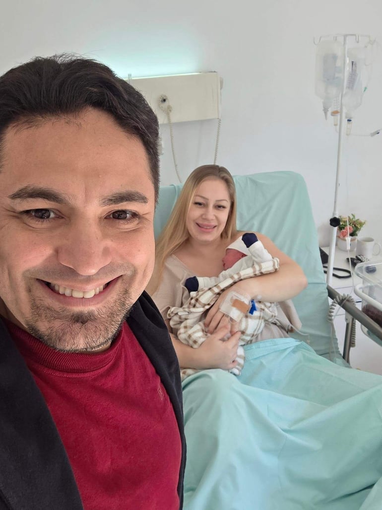 Hombre sonriente con camisa clara a la izquierda, y mujer en cama de hospital abrazando a su bebé envuelto en manta.