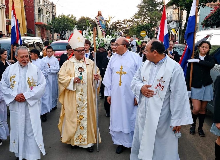 Hombre con báculo y vestimenta religiosa acompaña la procesión, donde jóvenes sostienen banderas en un entorno urbano.