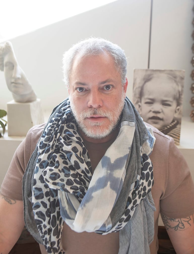 Hombre canoso con barba, camiseta marrón y bufandas gris y negro, sentado frente a una escultura y fotografía decorativa.