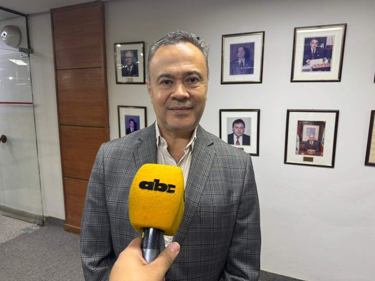 Humberto Blasco en blazer a cuadros con micrófono, rodeado de retratos en un ambiente institucional durante entrevista.