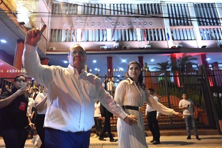 Camilo Pérez, con gafas y camisa blanca, sonríe y levanta un dedo índice, mientras una mujer con vestido blanco lo acompaña.