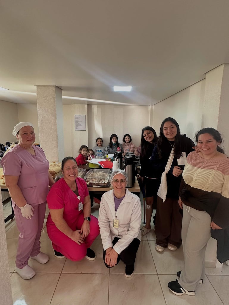 Cuatro mujeres en uniforme rosa y blanco, sonriendo y trabajando en un ambiente de cocina, rodeadas de más mujeres y niñas.