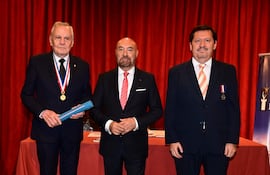 Mujer con abrigo oscuro y vestido claro junto a hombre de traje gris y corbata roja, en ambiente formal conversando.