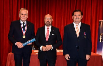 Mujer con abrigo oscuro y vestido claro junto a hombre de traje gris y corbata roja, en ambiente formal conversando.