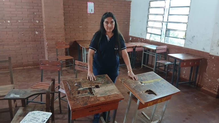 Mujer con camiseta azul inspecciona escritorios deteriorados en aula con mesas y sillas en mal estado.