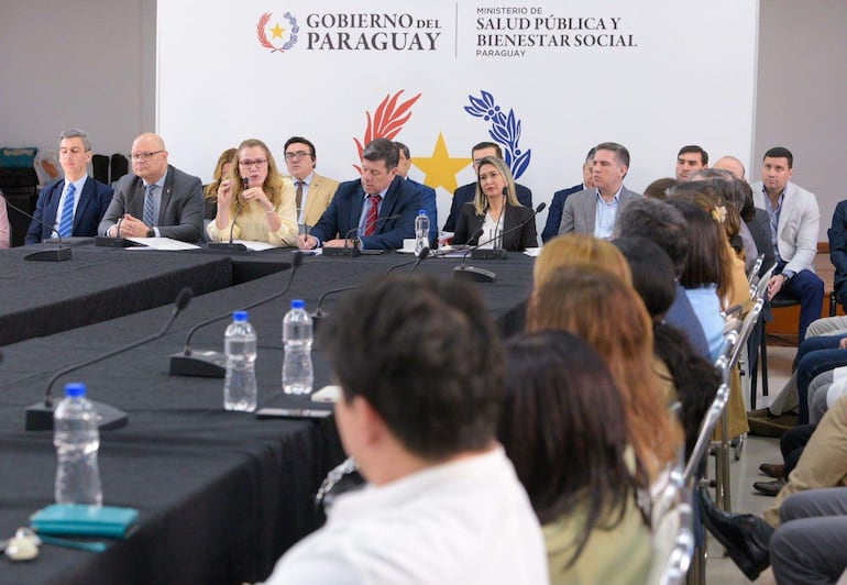 Mujer de cabello rubio habla en panel mientras asistentes prestan atención en un ambiente formal decorado con insignias del gobierno.