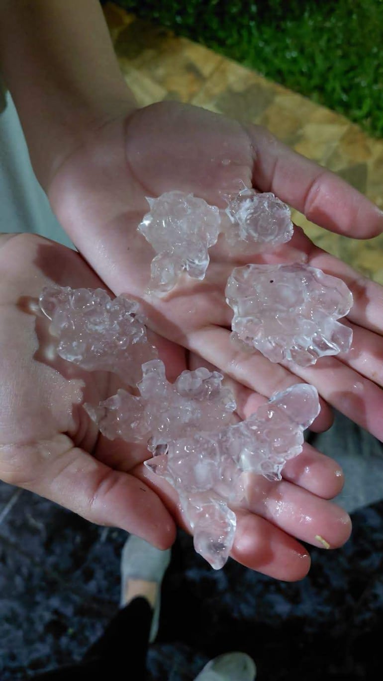 Manos sosteniendo trozos de granizo, mostrando su tamaño irregular y translúcido sobre un fondo de césped.