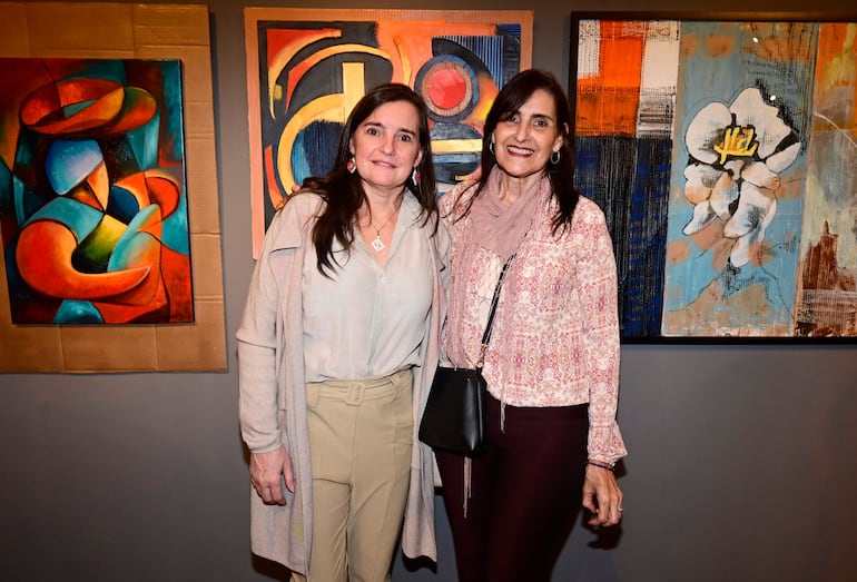 Dos mujeres sonríen frente a coloridas pinturas en una galería, una lleva blusa clara y pantalones beige, la otra blusa rosada y pantalones oscuros.