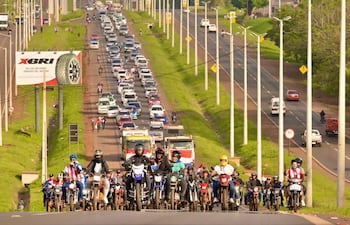 Motociclistas con banderas roja, blanca y azul lideran caravana, mientras vehículos permanecen detenidos en la carretera congestionada.