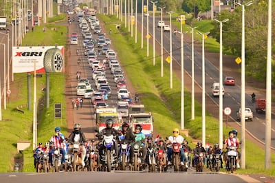 Motociclistas con banderas roja, blanca y azul lideran caravana, mientras vehículos permanecen detenidos en la carretera congestionada.