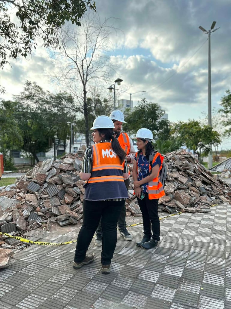 Tres personas en chalecos de seguridad conversan en un espacio público con escombros al fondo y clima nublado.