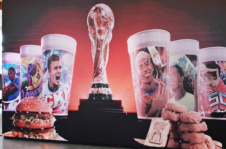 Trofeo de la Copa Mundial brilla en un montaje junto a una hamburguesa y vasos de colores en un entorno rojo.