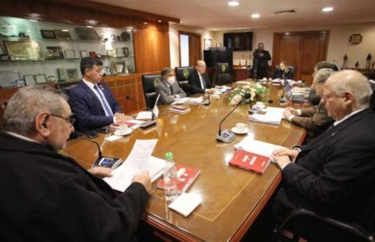 Víctor Ríos Ojeda en traje formal habla en reunión, rodeado de seis hombres atentos, con documentos y copas sobre la mesa.