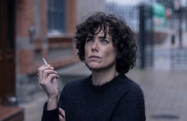 Patricia López Arnaiz con cabello rizado y suéter oscuro, sosteniendo un cigarrillo y mirando pensativa en un ambiente urbano.