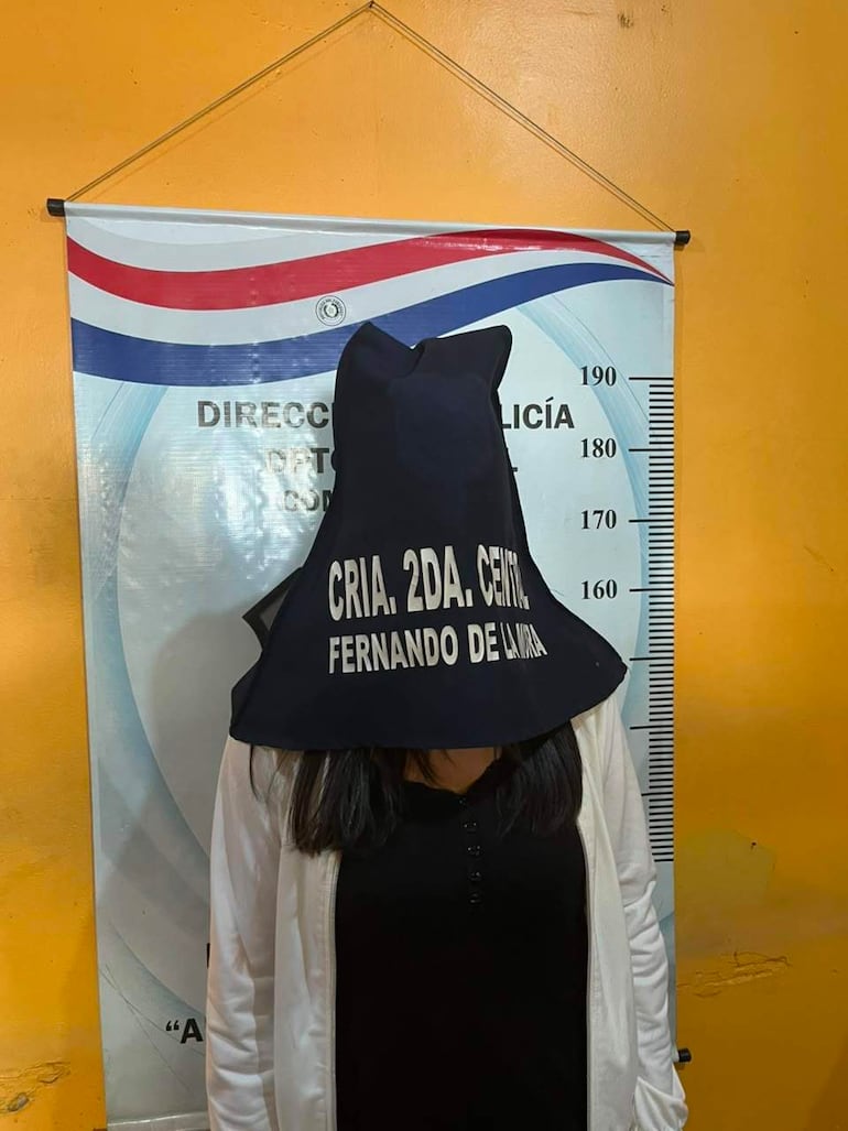 Mujer con chaqueta blanca y capucha negra en comisaría, rodeada de colores amarillos en un ambiente institucional.