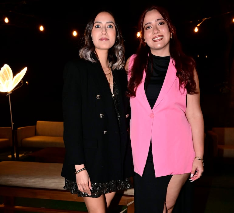 Mujeres sonrientes en evento nocturno; una lleva abrigo negro y vestido brillante, la otra en traje rosa claro.