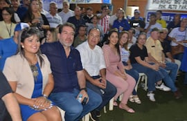 Grupo de personas en evento político, con María Portillo en blusa azul, sonriendo entre otros asistentes en un salón iluminado.