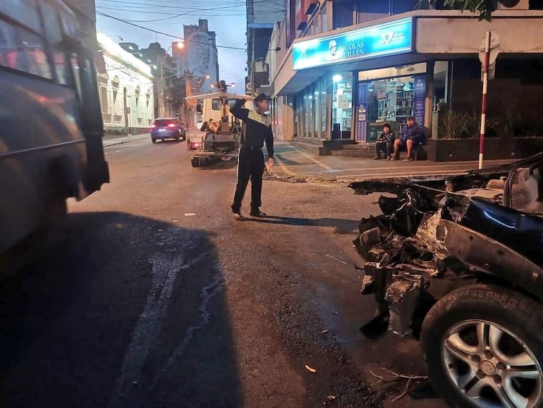 Hombre de pie dirigiendo el tráfico, autobús y vehículo dañado en una escena urbana. Dos hombres observan desde la acera.