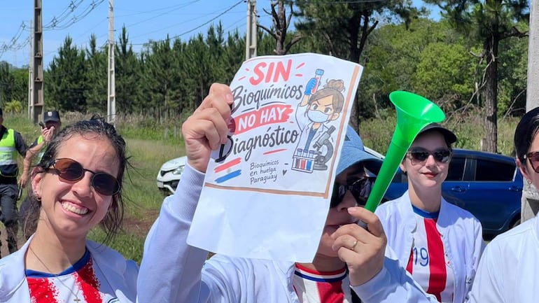 Tres mujeres sonrientes sostienen un cartel de protesta sobre bioquímicos, frente a un ambiente de manifestación al aire libre.