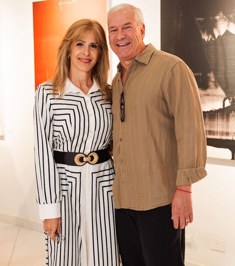 Mujer con camisa blanca a rayas y cinturón negro sonríe junto a hombre en camisa beige de rayas en galería con obras de arte de fondo.