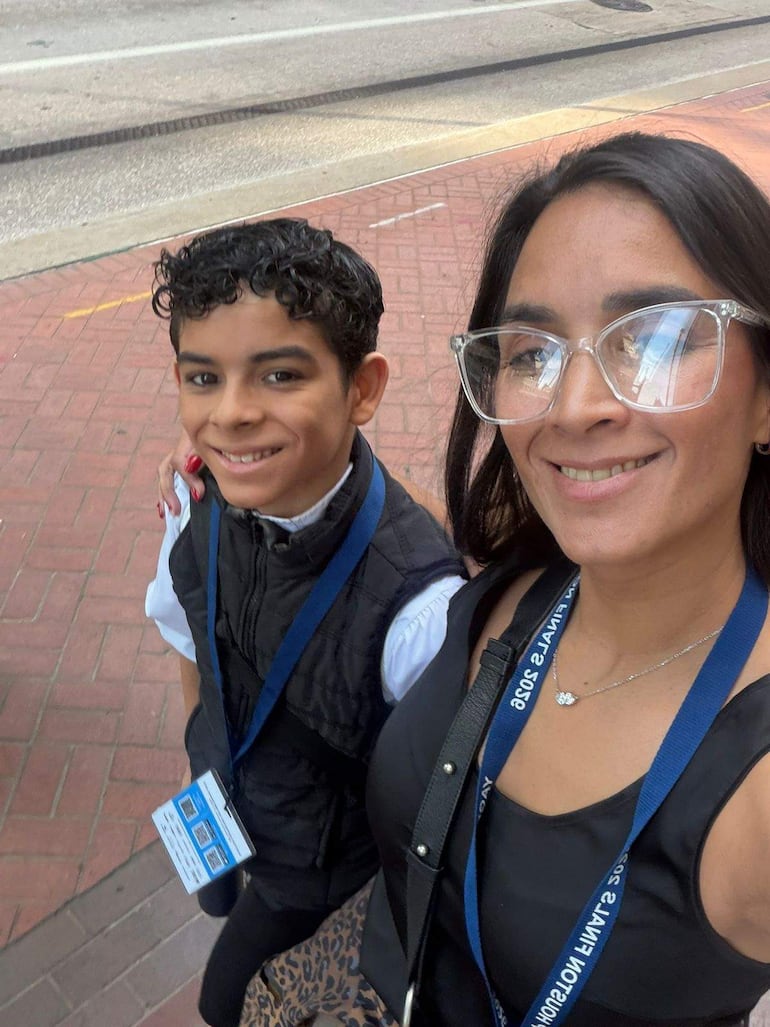 Mujer de cabello largo y gafas grandes sonríe junto a un niño con cabello rizado, ambos caminando en una acera con alegría.