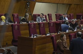 Interior del recinto legislativo, con personas en sillas rojas. Conversaciones privadas entre diputados y documentos sobre la mesa.