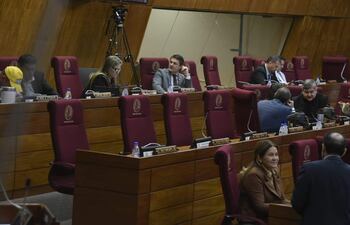 Interior del recinto legislativo, con personas en sillas rojas. Conversaciones privadas entre diputados y documentos sobre la mesa.