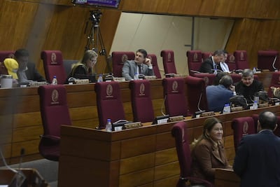 Interior del recinto legislativo, con personas en sillas rojas. Conversaciones privadas entre diputados y documentos sobre la mesa.