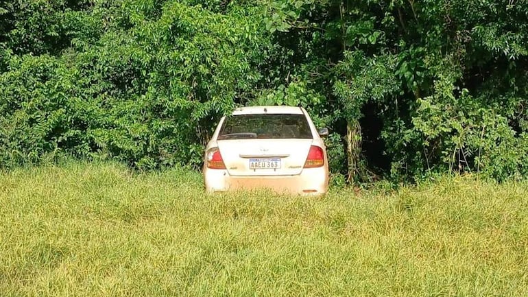Automóvil color claro abandonado en área boscosa, rodeado de hierba alta y sin personas visibles.