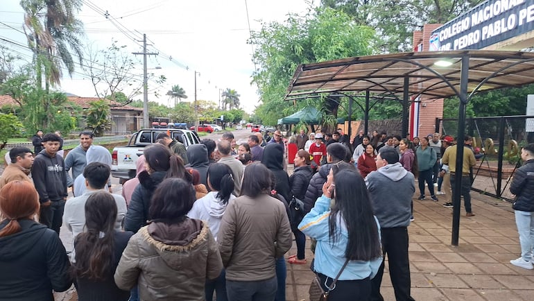 Grupo de electores en fila discutiendo frente al Colegio Nacional Dr. Pedro P. Peña, con un vehículo policial al fondo.