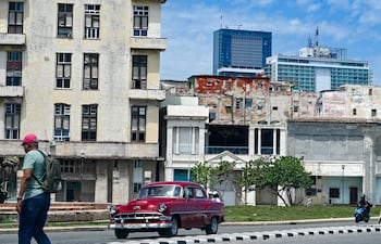 Un hombre camina por una calle mientras un auto pasa a su lado, con el hotel Iberostar Selection (C) y el hotel Habana Libre Melia (D) al fondo, en La Habana,