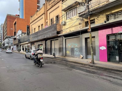 Motociclista con pasajero en la calle, rodeado de edificios y personas caminando; graffiti visible en una pared.