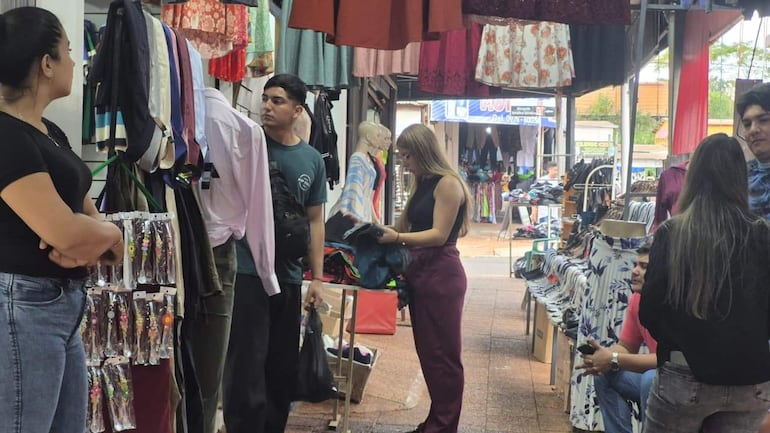 Mujer con cabello oscuro y camiseta negra observa a compradores en un mercado, con prendas colgadas alrededor.