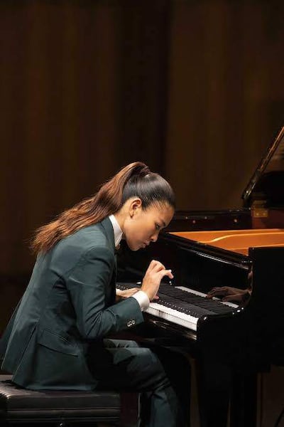 Músico Gile Bae, vestido con traje oscuro y cabello recogido, toca el piano con concentración en auditorio iluminado con luces cálidas.