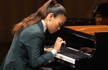 Músico Gile Bae, vestido con traje oscuro y cabello recogido, toca el piano con concentración en auditorio iluminado con luces cálidas.