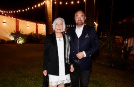 Pierre-Christian Soccoja en traje oscuro sonríe junto a una mujer con vestido blanco y abrigo negro, en un ambiente social iluminado.