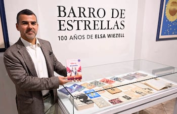 Hombre con saco marrón y camisa blanca sostiene un libro en un ambiente cultural, con cartel sobre Elsa Wiezell en el fondo.
