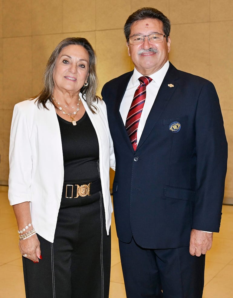 Mujer en blusa negra con abrigo blanco y falda negra sonríe junto a hombre en traje oscuro con corbata roja.