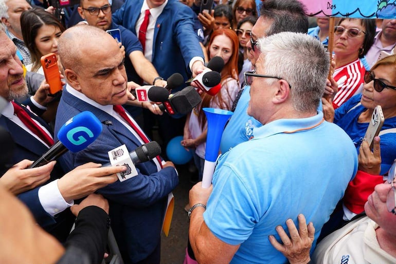 Basilio Gustavo Nuñez Gimenez en traje azul con corbata roja, sostiene un megáfono, mientras escucha a otro hombre en camiseta azul.