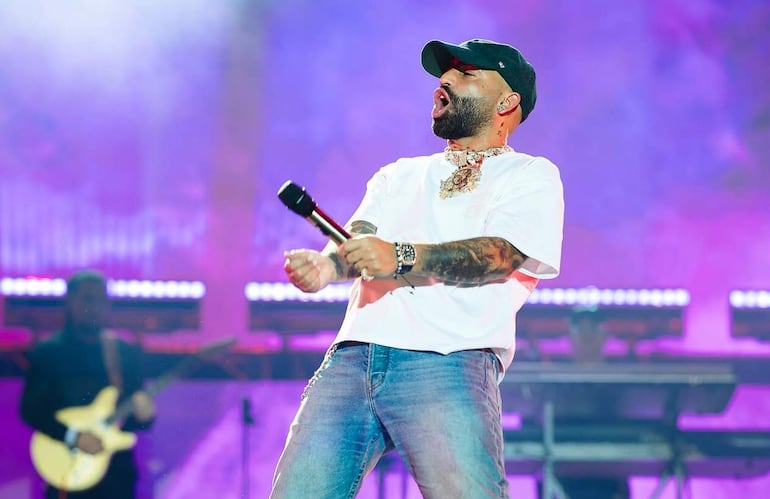 Hombre con camiseta blanca y jeans canta en el escenario sosteniendo un micrófono, mientras un guitarrista le acompaña en un ambiente festivo.