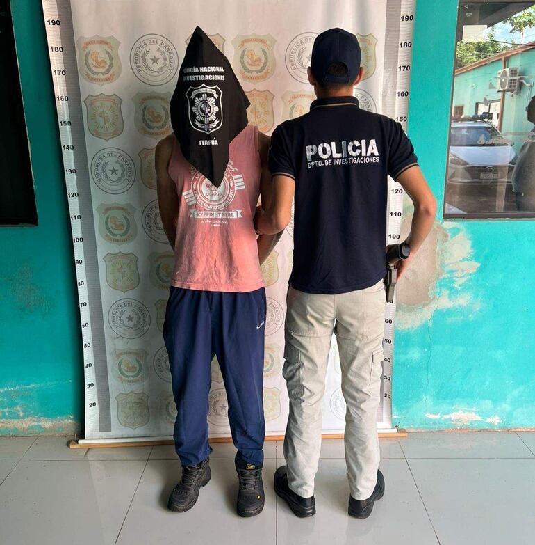 Joven con camiseta sin mangas rosa y bolsa negra en la cabeza, frente a un oficial de policía con arma, en el departamento de Investigaciones de Itapúa.